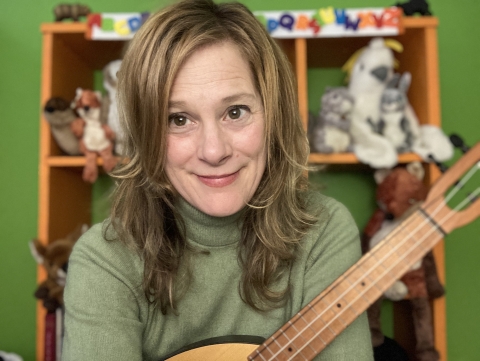 Sitting woman looking toward camera with a guitar-like instrument on her lap, puppets on shelves in the background