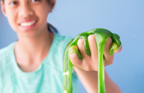 Smiling brown tween girl in background holding clear green slime in foreground