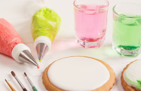 Round cookie with a circle of smooth white royal icing, with decorating bags , colored water, and paint brushes also on the table