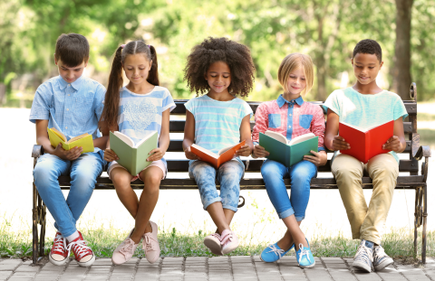 Five elementary age kids, boys and girls, sitting on a bench, each holding and reading a book