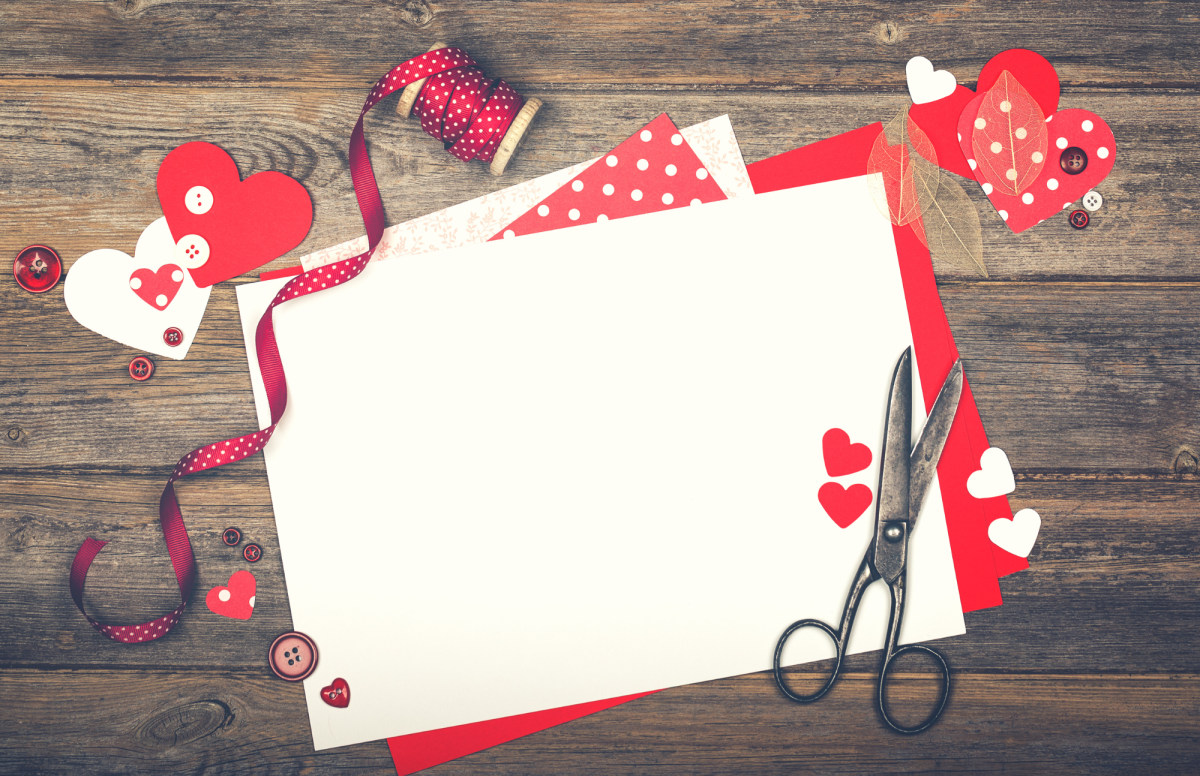 Red and white paper atop a wooden background, with cut out hearts and scissors