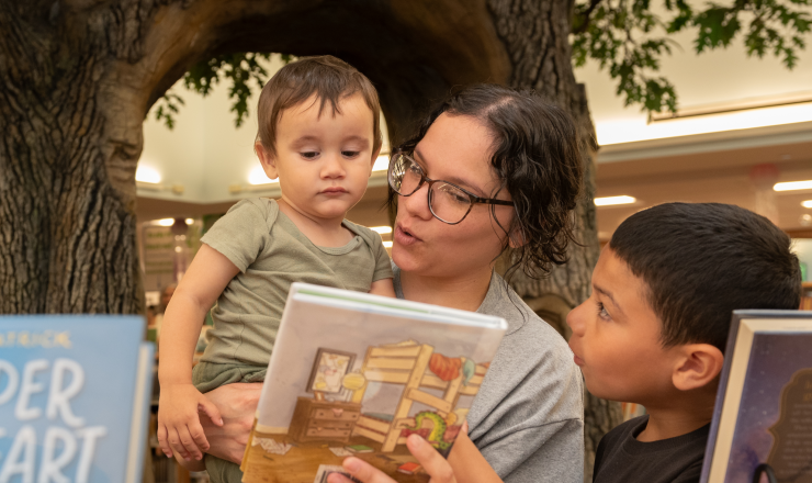 Mom holding young son showing him a book in the children's area with another young son standing beside her